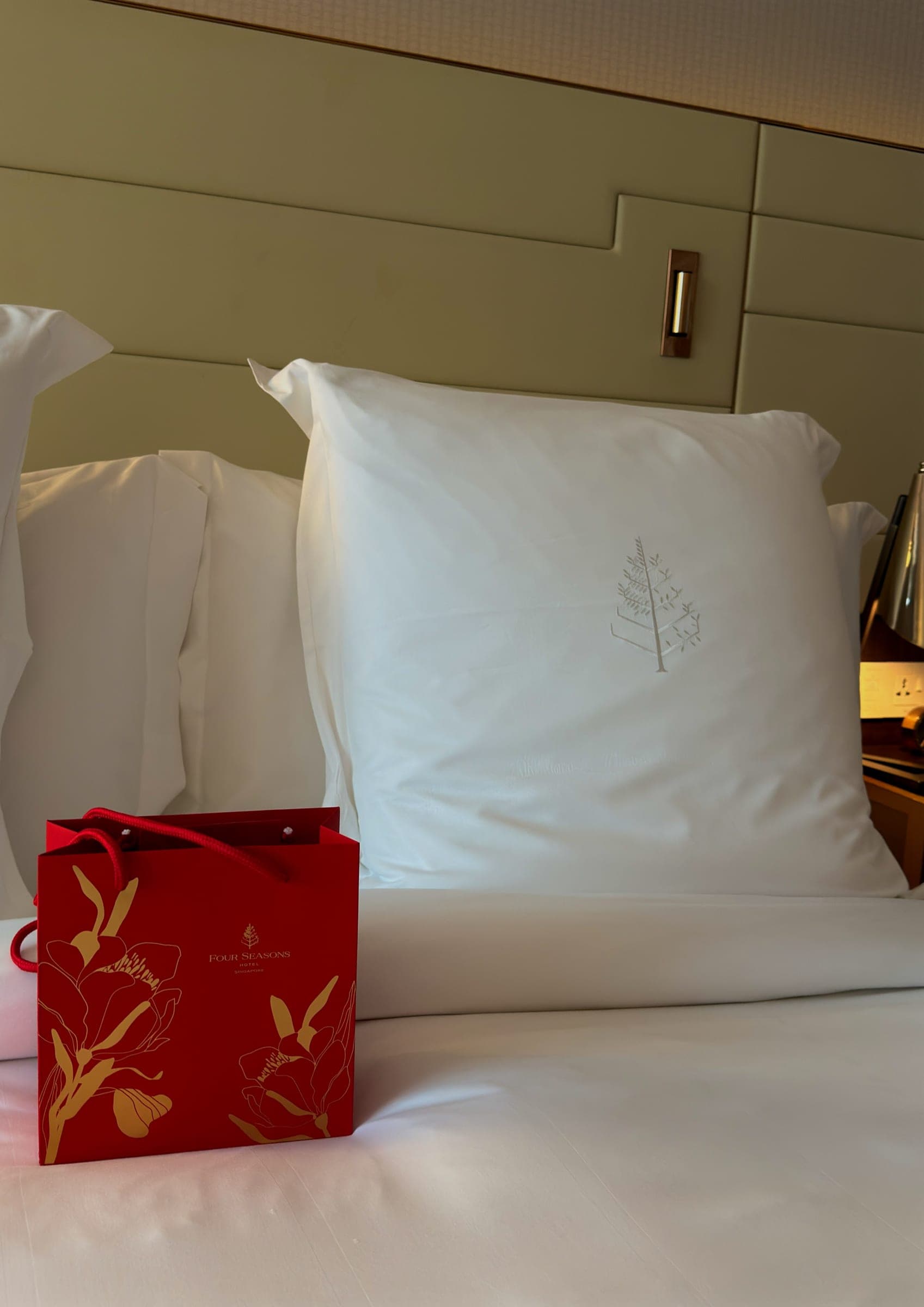 A crisp white pillow embroidered with the Four Seasons tree beside a red gift bag