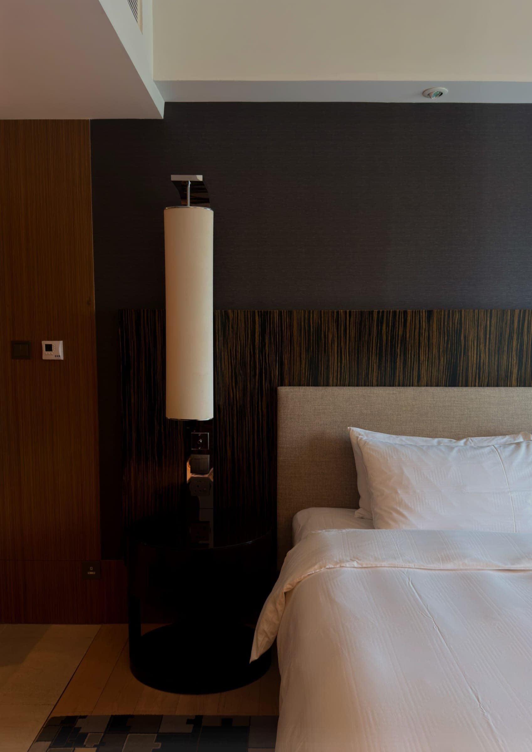 A bedside with a cylindrical lamp against a textured timber headboard