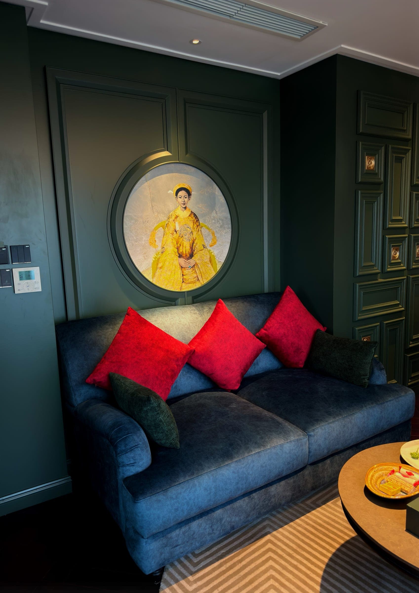 A blue velvet sofa with red cushions below a round portrait in a green panelled wall