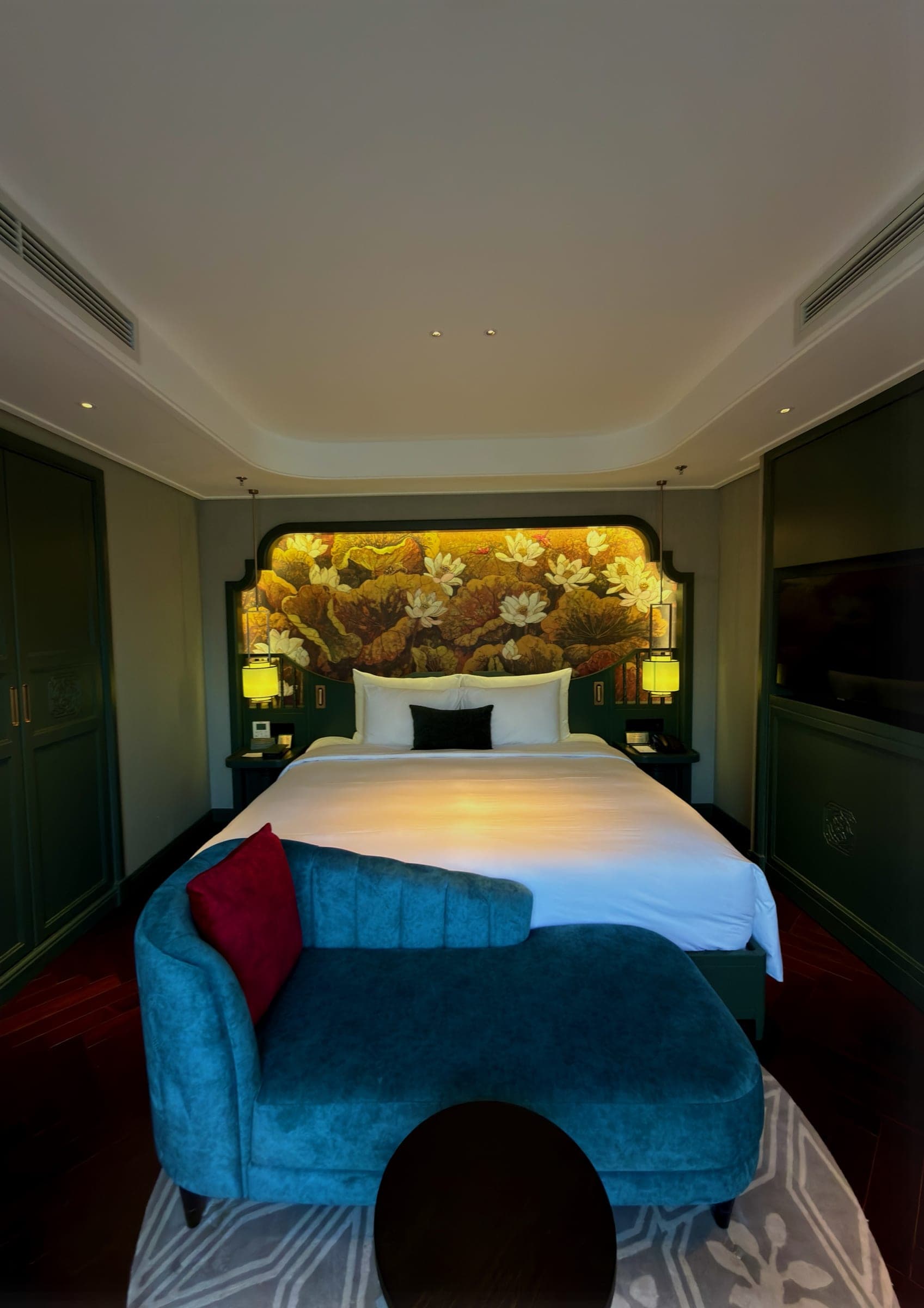 A bed beneath a painted lotus mural headboard with a blue velvet chaise at its foot
