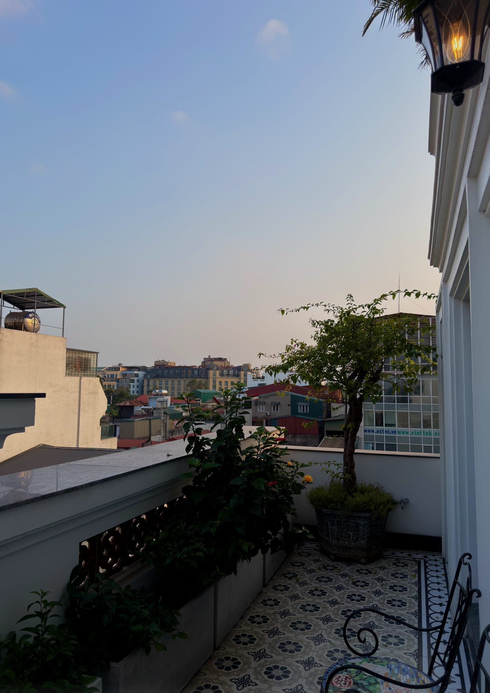 A tiled private balcony with plants and a bench above the Hanoi rooftops at dusk